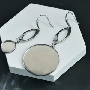 May include: A pair of silver earrings with a round pendant and an oval shaped dangle. The earrings have a hook closure.