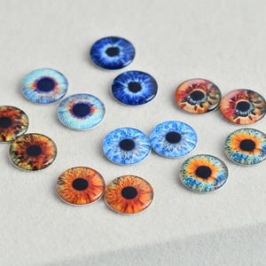 May include: A collection of glass eye cabochons in various colors, including blue, orange, and yellow. The irises feature detailed patterns and a dark pupil. These round, flat-backed glass eyes are suitable for crafts.
