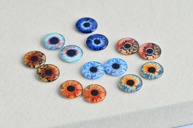 May include: A collection of glass eye cabochons in various colors, including blue, orange, and yellow. The irises feature detailed patterns and a dark pupil. These round, flat-backed glass eyes are suitable for crafts.