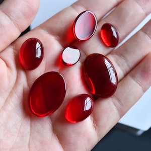 May include: Seven oval, translucent, deep red gemstones of varying sizes. The polished stones are displayed on a light background. The surfaces reflect light, creating highlights and shadows. Measurements are in centimetres.