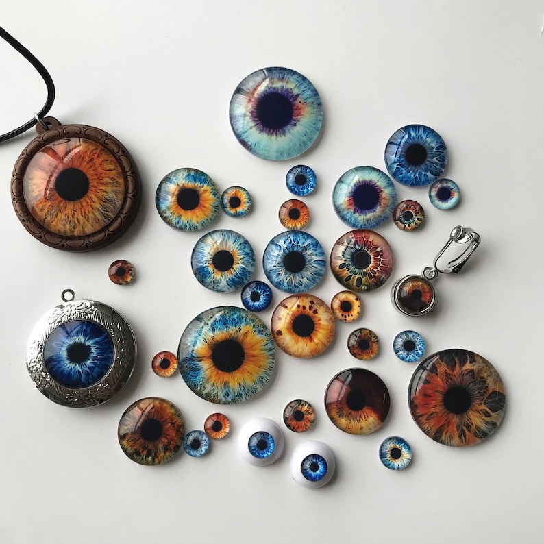 May include: A collection of eye-themed jewelry and craft supplies. Includes pendants, earrings, and glass cabochons with detailed iris designs in shades of blue, brown, and orange. Some pieces are set in metal or wood.