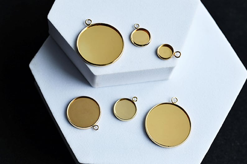 Puede incluir: Una colecci&oacute;n de colgantes redondos de color dorado, de varios tama&ntilde;os. Cada colgante tiene un peque&ntilde;o lazo en la parte superior para sujetarlo a una cadena u otro componente de joyer&iacute;a. Los colgantes se muestran sobre una superficie geom&eacute;trica blanca.