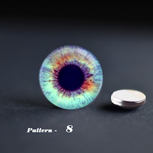 May include: A round, glass eye cabochon with a detailed iris pattern in shades of blue, green, orange, and purple. A small, silver-rimmed button is next to the cabochon. The text "Pattern - 8" is below the cabochon.
