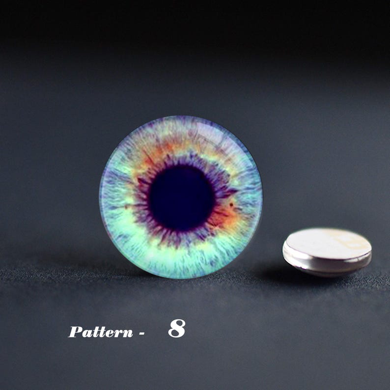 May include: A round, glass eye cabochon with a detailed iris pattern in shades of blue, green, orange, and purple. A small, silver-rimmed button is next to the cabochon. The text "Pattern - 8" is below the cabochon.