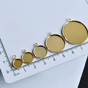 Puede incluir: Cinco bandejas redondas doradas para colgantes de varios tama&ntilde;os, cada una con un peque&ntilde;o lazo para sujetar una cadena. Las bandejas est&aacute;n dispuestas sobre una superficie blanca junto a una regla, la m&aacute;s grande mide unos 6,5 cm de di&aacute;metro.