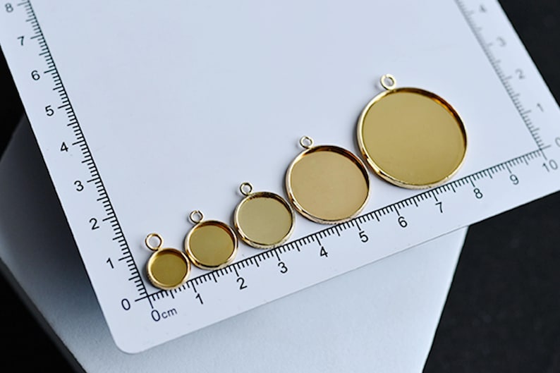 Puede incluir: Cinco bandejas redondas doradas para colgantes de varios tama&ntilde;os, cada una con un peque&ntilde;o lazo para sujetar una cadena. Las bandejas est&aacute;n dispuestas sobre una superficie blanca junto a una regla, la m&aacute;s grande mide unos 6,5 cm de di&aacute;metro.
