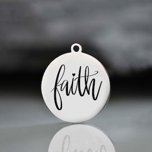 May include: A round, silver-colored pendant with the word "faith" in elegant black script. A small heart is placed above the "i". The pendant has a small loop at the top for hanging.