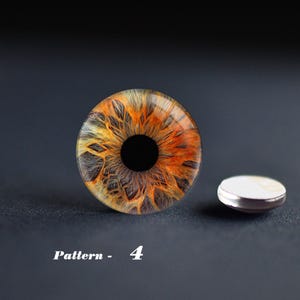 May include: A detailed, close-up of a glass eye with a striking orange, yellow, and black iris. The eye is round with a dark pupil. A small, silver-rimmed object sits beside it. The text "Pattern - 4" is below.