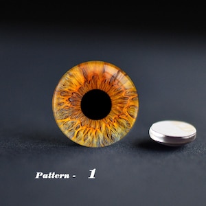 May include: A detailed, close-up of a glass eye with a vibrant orange and yellow iris and a black pupil. The eye is next to a small, round, silver-rimmed object. The text "Pattern - 1" is at the bottom.