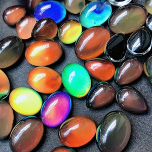 May include: A selection of oval-shaped, colour-changing stones in diverse hues. The stones transition from orange to green, blue, and purple, with some appearing brown or black. The polished stones have a smooth, glossy appearance, scattered across a dark surface.