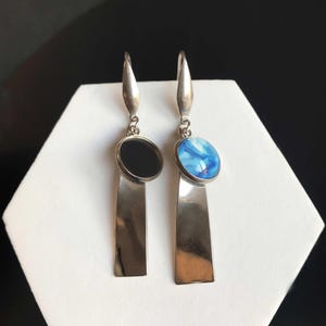 Pode incluir: Um par de brincos de prata com um design moderno. Um brinco apresenta uma pedra circular preta, enquanto o outro tem uma pedra marmorizada azul e branca. Ambos os brincos têm uma longa queda retangular de prata.