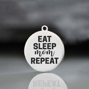 May include: A circular, silver-toned pendant with a small loop at the top. The pendant displays the words "EAT SLEEP mom REPEAT" in a bold, black font. The background is a blurred gradient of grey and black.