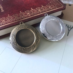 May include: Two round metal locket pendants with ornate floral designs. One pendant is silver and the other is bronze.