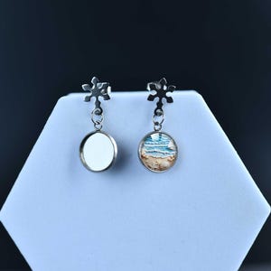 May include: A pair of silver-toned earrings. Each earring features a snowflake-shaped top and a circular pendant. One pendant is blank, while the other displays a beach scene with blue water and tan sand. The earrings are displayed on a white surface.
