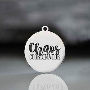 May include: A circular, silver-toned pendant with a small loop at the top. The pendant has the words "Chaos COORDINATOR" in a black, handwritten font. The pendant is reflective, with a blurred background.