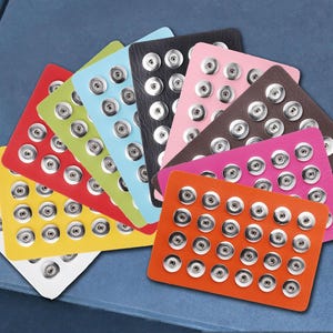 May include: A collection of rectangular cards, each featuring a grid of silver-toned metal snap fasteners. The cards are arranged in a fan shape and come in a variety of solid colors including white, yellow, red, lime green, light blue, black, pink, brown, magenta, and orange.