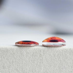 May include: Two glass cabochons with a realistic eye design. The cabochons feature a detailed iris in shades of orange and brown, with a black pupil. One cabochon is flat, and the other is domed. They are set against a neutral, textured background.