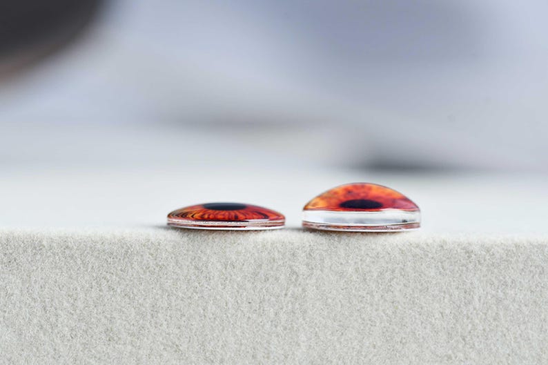 May include: Two glass cabochons with a realistic eye design. The cabochons feature a detailed iris in shades of orange and brown, with a black pupil. One cabochon is flat, and the other is domed. They are set against a neutral, textured background.