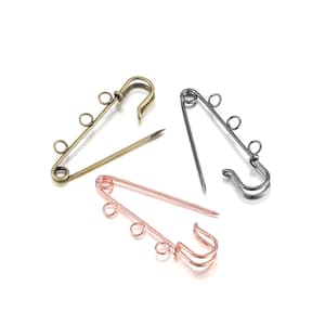 May include: Three metal safety pins in antique brass, gunmetal, and rose gold. Each pin has a looped head and three small loops for attaching charms or beads. The pins are arranged on a white surface.