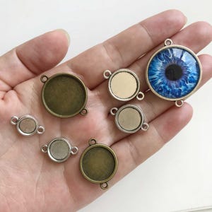 Pode incluir: Uma coleção de bandejas de pingente redondas em vários tamanhos e acabamentos, incluindo bronze antigo e prata. Um pingente apresenta um design detalhado de olho azul. Cada bandeja tem laços para prender a uma corrente ou outro componente de joalheria.