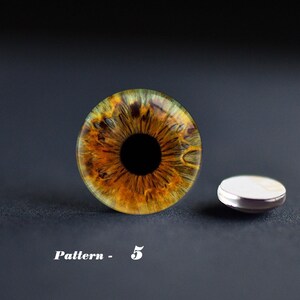 May include: A detailed close-up of an eye with a brown and yellow iris and a black pupil. The eye is next to a small, round, silver-rimmed object. The text "Pattern - 5" is below the eye.