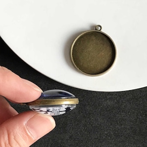 Puede incluir: Una base de colgante redonda con acabado bronce se encuentra junto a un colgante con una cúpula de cristal azul. El colgante tiene un diseño en blanco y negro con la palabra "HOPE". El colgante está sostenido por una mano.