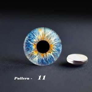 May include: A detailed close-up of a blue and yellow eye pattern, with a black pupil. A small, round, silver-rimmed object is next to the eye design. The text "Pattern - 11" is below the eye design.