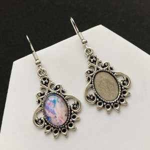 Puede incluir: Un par de pendientes de plata con diseños de filigrana ornamentados. Un pendiente presenta un cabujón de vidrio ovalado transparente, mientras que el otro pendiente tiene un diseño abstracto colorido.