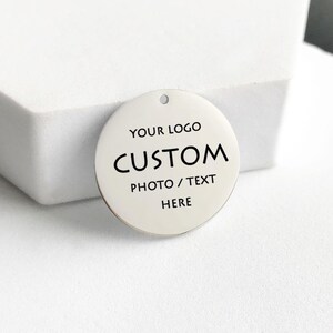 Peut inclure: Un charm rond argenté avec un petit trou en haut. Il porte les inscriptions en noir : "YOUR LOGO", "CUSTOM", et "PHOTO / TEXT HERE". Idéal pour la personnalisation.