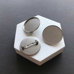 Pode incluir: Três bases redondas de metal prateado para broche. Duas bases são planas e uma base tem um pino.