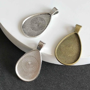 Puede incluir: Tres bandejas de colgantes en forma de lágrima plateadas y bronce con un patrón decorativo. Las bandejas están diseñadas para contener un pequeño camafeo u otro objeto decorativo.