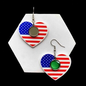 May include: A pair of heart-shaped earrings with an American flag design. Each earring features a silver hook and a round, clear glass cabochon in the center. One cabochon is silver and the other is green.
