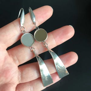 May include: A pair of silver-toned dangle earrings. Each earring features a circular element, a small connecting ring, and a long, triangular pendant. The earrings are displayed against a dark background.