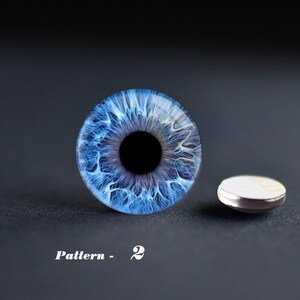 May include: A detailed image of a blue eye with intricate patterns and a black pupil. Next to it is a small, round, silver-rimmed object with a white top. The text "Pattern - 2" is displayed below the eye.