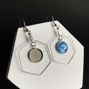 May include: A pair of silver hexagonal hoop earrings with a round pendant hanging from each hoop. One pendant is clear glass and the other is blue with a white swirl pattern.