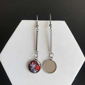 Puede incluir: Un par de pendientes colgantes plateados. Cada pendiente tiene una barra larga y delgada suspendida de un gancho. Uno presenta un diseño circular rojo y blanco, el otro un círculo gris claro en blanco. Miden unos 6 cm de largo.