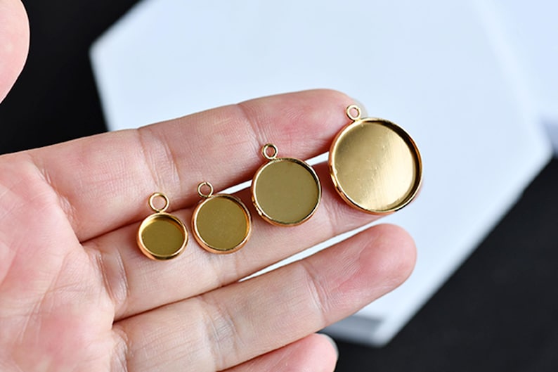 Puede incluir: Cuatro bandejas redondas doradas para colgantes de diferentes tama&ntilde;os, cada una con un peque&ntilde;o lazo en la parte superior para sujetar una cadena o cord&oacute;n. Las bandejas se sostienen en una mano, mostrando sus diferentes di&aacute;metros. Las bandejas tienen una superficie brillante y reflectante.