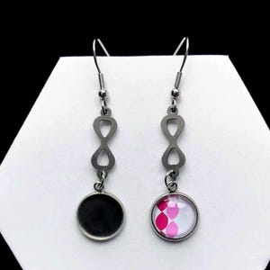 May include: A pair of silver-toned dangle earrings. Each earring features an infinity symbol design and a round pendant. One pendant is black, the other has a pink and white geometric pattern. The earrings hang from a fish hook.