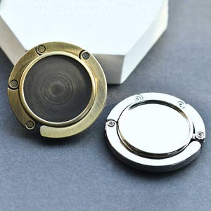 May include: Two circular folding bag hangers, one with a brushed antique brass finish and a dark center, and the other with a polished silver-tone finish and a reflective center. Both items feature a hinged design with small circular accents.