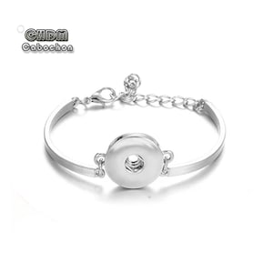 May include: A silver-toned metal bracelet featuring a central circular snap button base for interchangeable inserts. The bracelet includes a thin, smooth band, a lobster claw clasp, and an adjustable chain link extension. Text reads CHDM Cabochon.