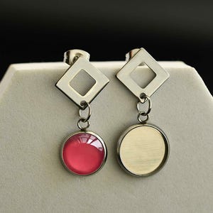 May include: A pair of silver earrings with a square design and a dangling round charm. One charm is a clear glass cabochon, the other is a pink glass cabochon.