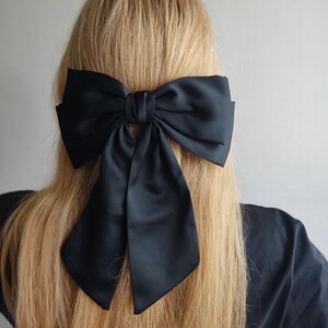 Könnte beinhalten: Ein großer schwarzer Satin-Haarschmuck mit Schleife. Die Schleife ist in der Mitte gebunden und hat lange, fließende Enden. Der Haarschmuck befindet sich vor dem Hintergrund von langem, blondem Haar.