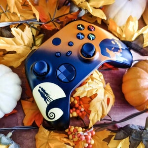May include: A black and orange Xbox controller with a Halloween-themed design featuring a silhouette of Jack Skellington from The Nightmare Before Christmas. The controller is surrounded by fall leaves and pumpkins.