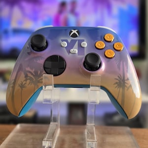 Op de afbeelding: Een draadloze videogamecontroller met een zonsondergang-kleurverloop van blauw, paars en oranje met zwarte palmbomsilhouetten. Het bevat goudkleurige actieknoppen, zwarte joysticks, een richtingspad en de tekst VI.