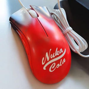 May include: A red computer mouse with the text "Nuka Cola" in white script. The top of the mouse has a gradient design with stars. The mouse has a white scroll wheel and a white cord.