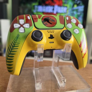 May include: A custom video game controller with a Jurassic Park theme. The controller has a yellow base with green and red accents, a fern leaf design, and the Jurassic Park logo. The buttons are white and black.