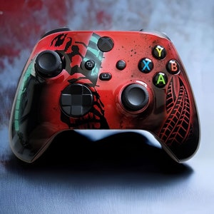 Może przedstawiać: Czerwono-czarny kontroler Xbox z niestandardowym wzorem przedstawiającym skorpiona i wzór bieżnika opony. Kontroler ma zielone, żółte i niebieskie przyciski.