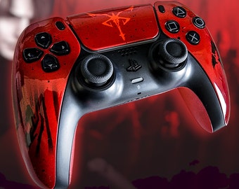 Puede incluir: Un mando PlayStation 5 DualSense rojo y negro con un diseño personalizado que presenta un símbolo en blanco y negro. El mando tiene una superficie texturizada y un logotipo de PlayStation negro.