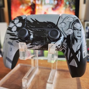 Könnte beinhalten: Ein schwarz-weißer PlayStation 5-Controller mit einer stilisierten Grafik eines Charakters. Der Controller steht auf einem durchsichtigen Plastikständer.