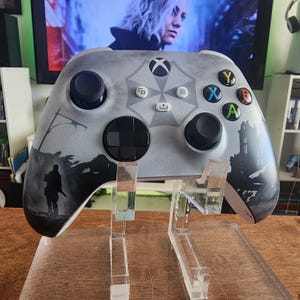 May include: Custom-designed Xbox controller in shades of gray, featuring a detailed silhouette scene and the Umbrella Corporation logo. The controller has black joysticks and buttons with colored accents, displayed on a clear stand.